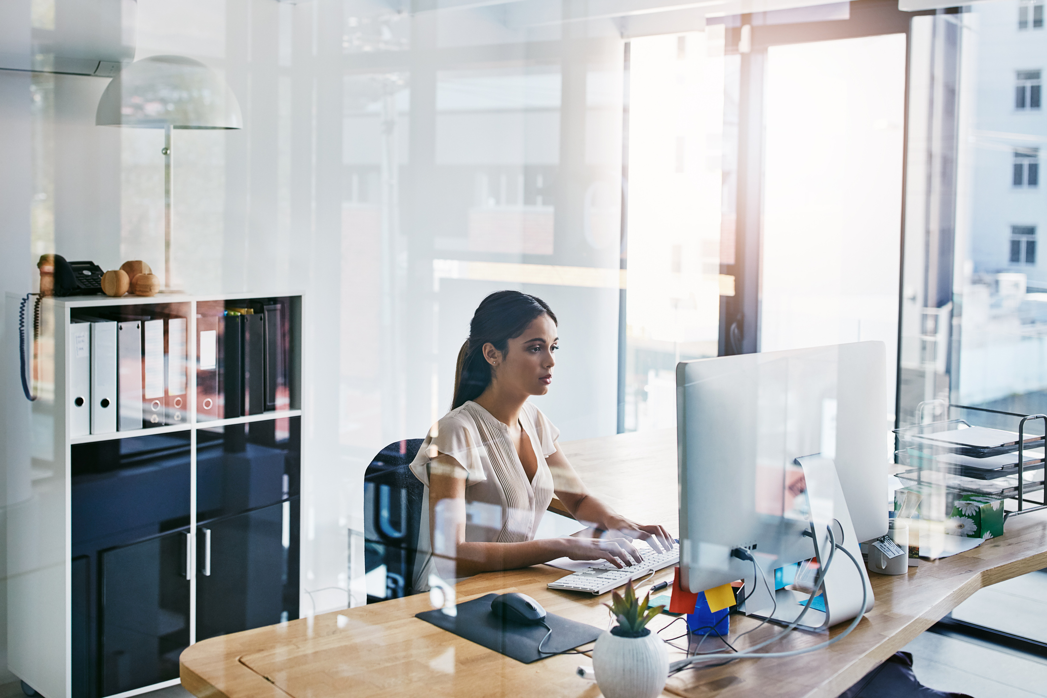 A woman in an office with clear walls is sitting at a desk and working on a computer