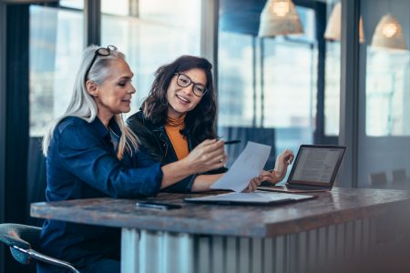 two female colleagues standing next to each other at a high counter in an office with a laptop and a sheet of paper working on ACH payment processing account.