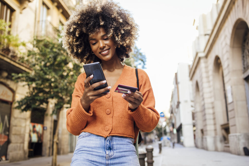 African-American woman holding both her smartphone and credit card along the street